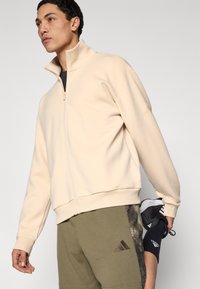 Μπεζ ζακέτα με φερμουάρ από απαλό ύφασμα, με ψηλό γιακά και ελαστικά τελειώματα στα μανίκια, συνδυασμένη με χακί σορτς που διαθέτουν λογότυπο και σχέδιο στο πλάι.