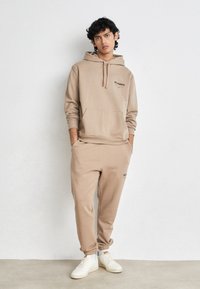 Beige kapucnis pulóver elülső zsebbel és fekete logóval, illeszkedő, tapered fazonú melegítőnadrággal párosítva. A modellen fehér sportcipő van. Egyszerű háttér.