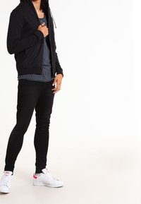 Felpa nera con zip, indossata sopra una maglietta grigia, abbinata a pantaloni neri slim-fit e sneakers bianche con accenti rossi.