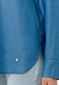 Jasnoniebieska koszula dżinsowa z zaokrąglonym dołem, z małym białym guzikiem na dole. Gładka faktura i długie rękawy z guzikowymi mankietami.