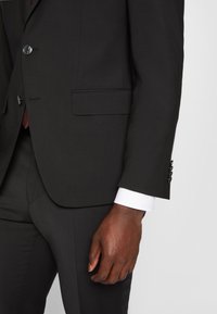 Zwarte blazer met pieklapellen, sluiting met één knop en een zijzak. Heeft een donkere, gestructureerde stof en een contrasterende witte manchet.