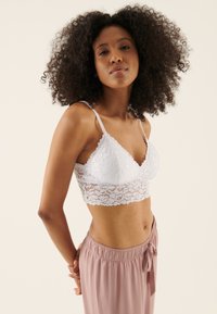 White lace bralette featuring a triangular shape, delicate floral pattern, and adjustable straps; worn with pink relaxed trousers.