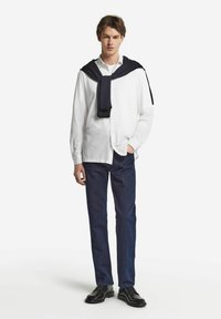 Camicia bianca con bottoni, maglione blu navy appoggiato, pantaloni in denim scuro, scarpe nere. Linee pulite, texture lisce e una palette di colori neutri.