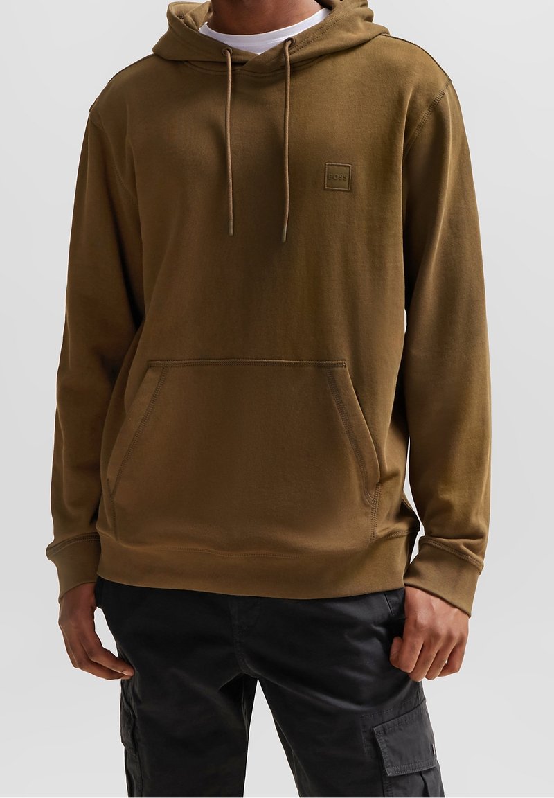 Homme portant un sweat à capuche marron avec poche frontale et cordons, associé à un pantalon cargo noir, debout devant un fond uni.