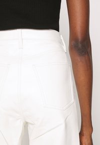 Rückansicht einer Person, die hochgeschnittene weiße Hosen mit großen Gesäßtaschen und ein schwarzes Rippenshirt trägt. Der Arm hängt entspannt an der Seite.