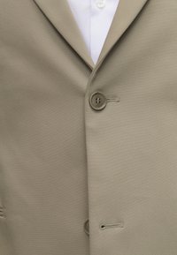 Gros plan d'un blazer beige boutonné sur une chemise blanche avec col et détails des boutons visibles.