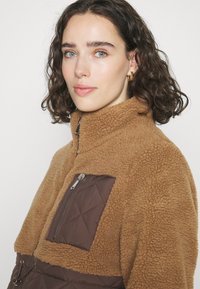 Καφέ φλις πουλόβερ με ψηλό λαιμό, που διαθέτει τσέπη με καπιτονέ σχέδιο διαμαντιών και ομαλό φερμουάρ. Μαλακή υφή με χαλαρό σχέδιο.