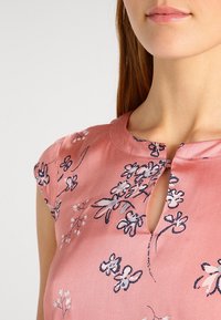Kurze pinke Bluse mit kurzen Ärmeln, die mit blauer und weißer Blumenstickerei verziert ist und über ein Schlitz-Ausschnittdetail verfügt. Glatte Stofftextur.