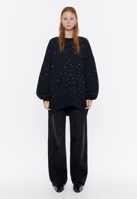 Sudadera negra de gran tamaño con puntos multicolores, mangas largas y dobladillo curvado, combinada con pantalones negros de pierna ancha y zapatos de punta.