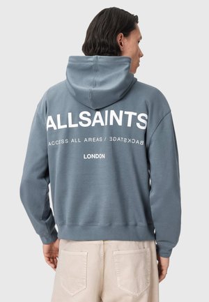 Osoba nosząca niebieską bluzę z kapturem z napisami "ALLSAINTS", "ACCESS ALL AREAS / BACKSTAGE" i "LONDON" na plecach, w połączeniu z jasnobeżowymi spodniami.