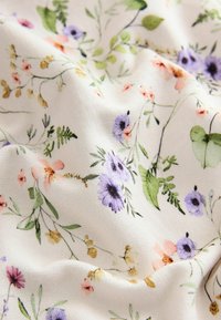 Vit tyg med ett blommönster som har lila, rosa och gröna blommor. Texturen är mjuk med subtila veck och skrynklor.