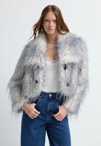 Faux-Fur-Jacke mit einem grau-weißen Muster, das eine zottelige Textur aufweist, im Doppelreiher-Design und zwei schwarzen Knöpfen. Kombiniert mit blauen Jeans.