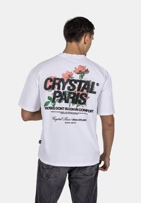 Bílý tričko s multikolorovým květinovým designem na zadní straně a černým textem. Krátké rukávy, kulatý výstřih a volný střih.