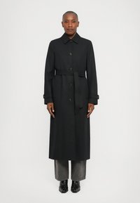 Melns garš mēteli ar pogu priekšpusi, plašu apkakli un jostu viduklī. Iezīmējas ar izteiksmīgām pogām un smalku tekstūru. Valkāts virs pelēkiem biksēm un zābakiem.