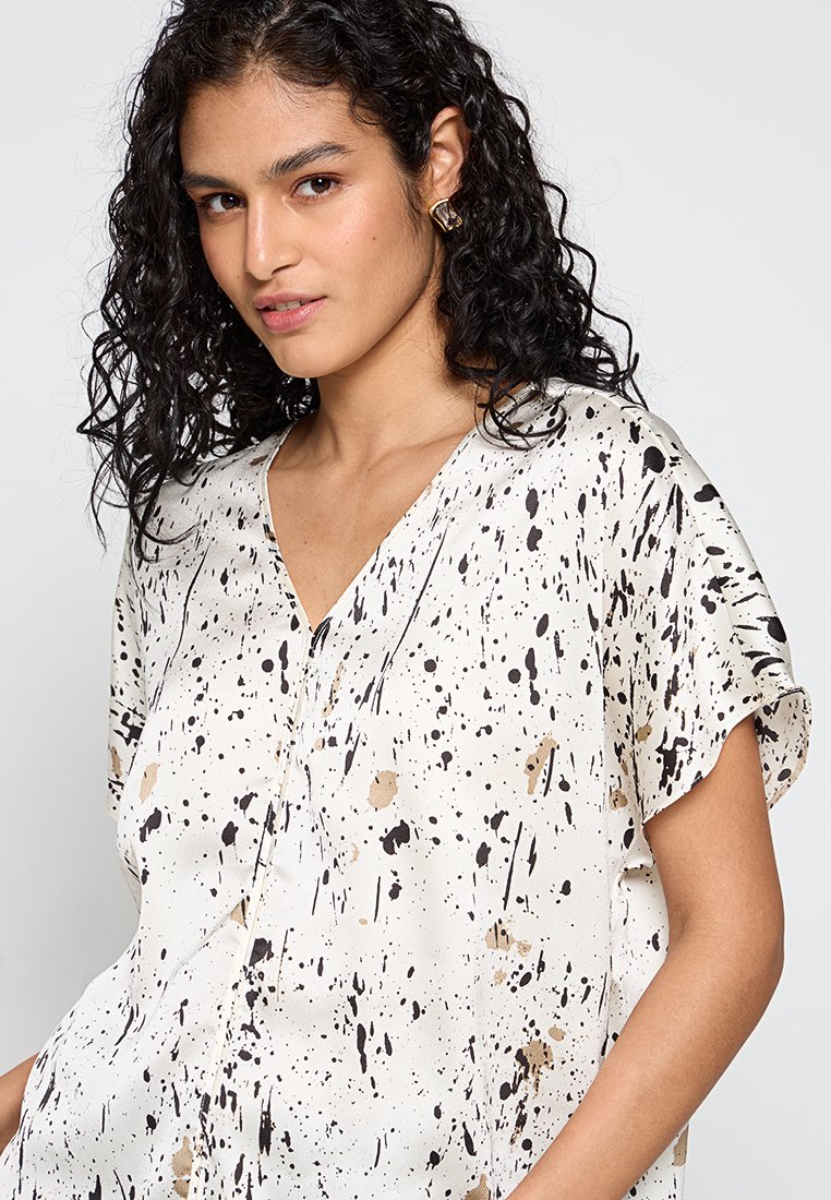 Lichte, crèmekleurige blouse met zwarte en gouden spetters. Heeft een V-hals, korte mouwen en een comfortabele pasvorm.