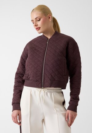 Frau mit blondem Pferdeschwanz, die eine gesteppte dunkelviolette Reißverschlussjacke und weiße Kordelzughose mit dunklen Seitenstreifen trägt.