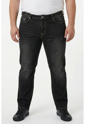 Mann trägt ausgewaschene schwarze Jeans, schwarze Sneakers und ein weißes Shirt, steht vor einem einfachen weißen Hintergrund.