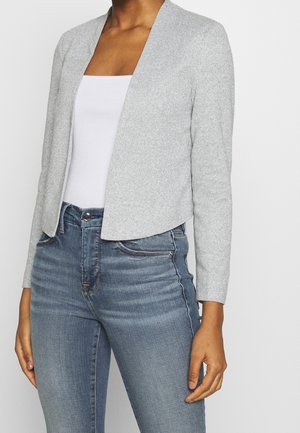 Vrouw draagt een lichtgrijze open cardigan, wit geribd hemdje en hooggetailleerde blauwe spijkerbroek tegen een effen achtergrond.
