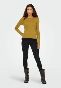 Sennepsfarvet ribbet langærmet top, tætsiddende design, parret med sorte skinny jeans og brune ankelstøvler med spænde detaljer.