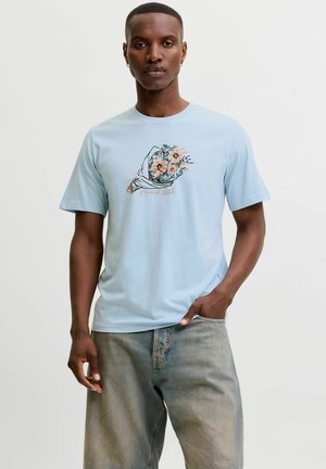 Mand iført lyseblå t-shirt med en illustration af en blomsterbuket og lysegrå jeans, står med den ene hånd i lommen.