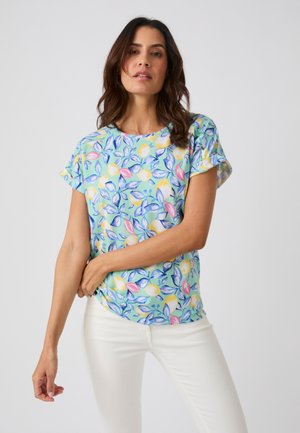 Donna che indossa una maglietta a maniche corte azzurro pastello con motivi floreali e foglie gialle e rosa e pantaloni bianchi, posando su uno sfondo semplice.