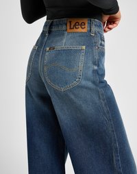 Back view of person wearing blue Lee brand jeans with stitched pockets and brown leather Lee logo patch on the waistband.