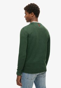 Młody mężczyzna zwrócony bokiem, ubrany w ciemnozieloną koszulę z długim rękawem i jasnoniebieskie dżinsy na tle jednolitej białej ściany.