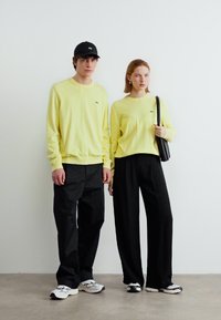 Νεαρός άνδρας και γυναίκα φορούν ταιριαστά ανοιχτού κίτρινου χρώματος φούτερ, μαύρο παντελόνι και αθλητικά παπούτσια, στέκονται δίπλα-δίπλα μπροστά σε λευκό τοίχο.