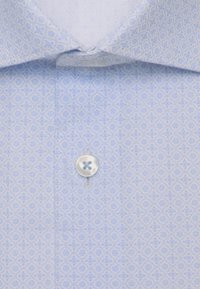 Chemise bleu clair à petit motif géométrique répétitif avec un seul bouton blanc fermé sous le col.