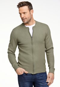 Maglione verde con zip, colletto e polsini a coste, realizzato in tessuto a maglia. Indossato sopra una camicia bianca e abbinato a jeans di denim scuri.