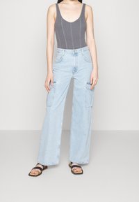 Žena v šedém žebrovaném tílku, světle modrých širokých kapsáčích a černých sandálech se čtveratou špičkou stojící na jednobarevném pozadí.