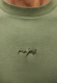 Olivzaļa sporta krekla ar ribotām apkaklēm un smalku uzšuvu "mo'pd" uz krūtīm. Gluda auduma tekstūra.