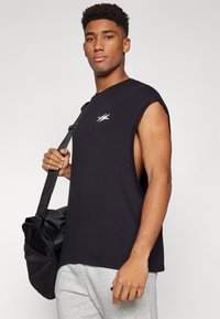 Camiseta sin mangas negra con escote redondo, que presenta un logo blanco. Combinada con pantalones de chándal grises y una bolsa de gimnasio negra.