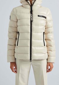 Beige dunjakke med polstret design, sort lynlås, forlommer og logoaccenter. Glat, blank tekstur med hætte.