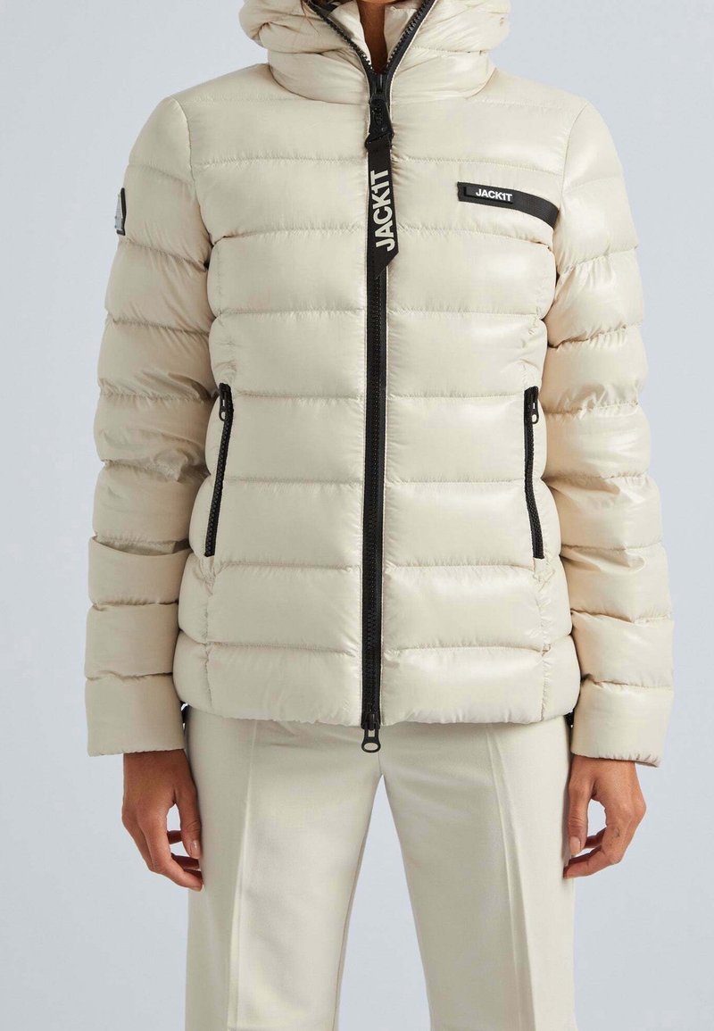 Beige dunjakke med polstret design, sort lynlås, forlommer og logoaccenter. Glat, blank tekstur med hætte.