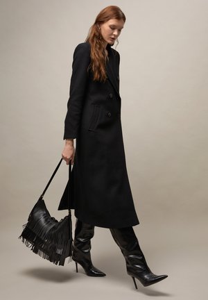 Mujer caminando con un abrigo largo negro de doble botonadura, botas negras de cuero con tacón alto y llevando un bolso negro con flecos.