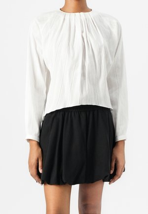 Femme portant un chemisier blanc plissé à manches longues et une jupe noire taille haute et coupe décontractée sur un fond uni.