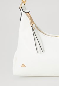 Sac à main en cuir blanc avec une fermeture éclair dorée, des accents noirs et un petit logo triangulaire doré. Finition texturée avec une forme arrondie.