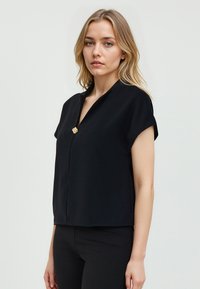 Top nero a maniche corte con leggero scollo a V, caratterizzato da un'accento floreale dorato al centro e un fit rilassato. Texture morbida e liscia.