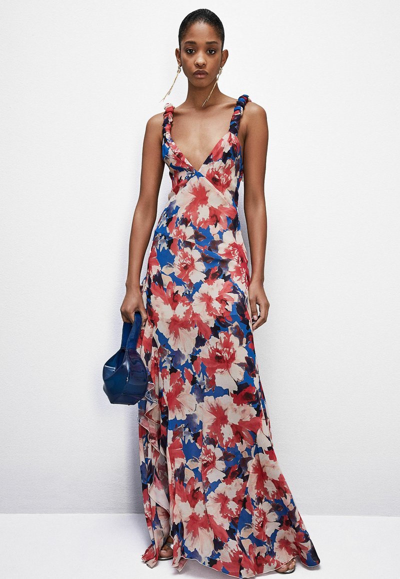 Abito maxi floreale con scollatura a V profonda, caratterizzato da colori blu, rosso e crema, con spalline arricciate. Abbinato a una borsa blu.