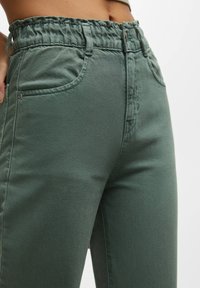 Pantalon en denim vert avec une taille élastique, deux poches avant et une légère texture. Présente une coupe ajustée et un design décontracté.