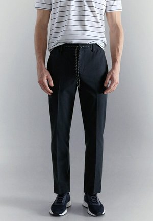 Hombre con pantalones negros a medida con cinturón de cuerda, zapatillas azul marino y camisa blanca de manga corta con rayas horizontales negras.