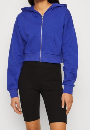 Afgeknotte hoodie in levendig blauw, met een ritssluiting aan de voorkant en een grote zak, gecombineerd met zwarte geribbelde fietsshorts.