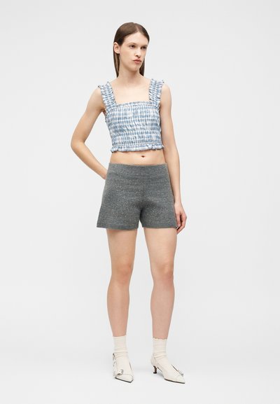 Νεαρή γυναίκα που φοράει μια μπλε και λευκή καρό κοντομάνικη μπλούζα, γκρι πλεκτές σορτς, λευκές κάλτσες και λευκά παπούτσια με μυτερή μύτη, στέκεται μπροστά σε απλό φόντο.