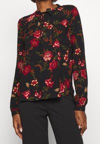 Schwarze Bluse mit Blumenmuster und langen Ärmeln, verziert mit roten und rosa Blumen, mit einer Schleifen-Nackenpartie und entspannter Passform. Weicher Stoff mit glatter Textur.