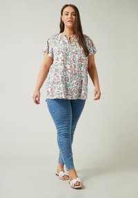Blumenbluse mit kurzen Ärmeln und Schlüssel-Ausschnitt, cremefarbener Hintergrund, mehrfarbiges Blumenmuster; kombiniert mit blauen Slim-Fit-Jeans und weißen Sandalen.
