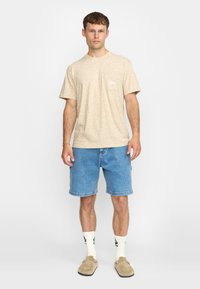 Beige textuur T-shirt met korte mouwen en een klein logo patch, gecombineerd met blauwe denim shorts en crèmekleurige sokken, afgewerkt met tan instappers.