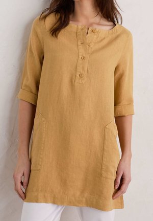 Kobieta ubrana w lnianą tunikę w kolorze musztardowożółtym z rękawami do łokcia, zapinaną na guziki i z naszywanymi kieszeniami z przodu, w połączeniu z białymi spodniami.