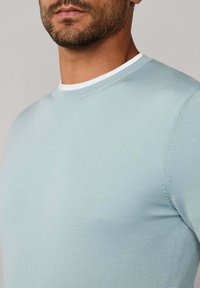 Detailní záběr na muže v světle modrém svetru s kulatým výstřihem přes bílou spodní košili, kde jsou vidět detaily jeho krku a vousů.