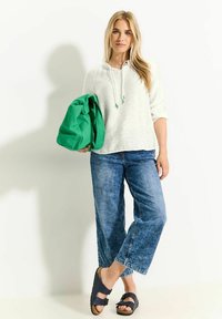 Haut à capuche blanc à manches longues avec un cordon, jean bleu large, et sandales foncées. Tient un sac vert. Tenue simple et décontractée.